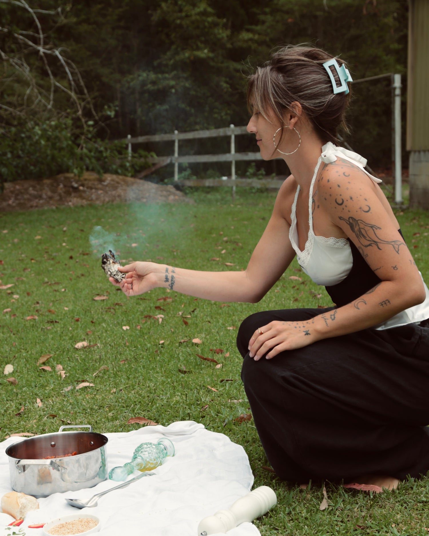 Woman squatting outdoors with a pot and other items on a blanket, surrounded by greenery.