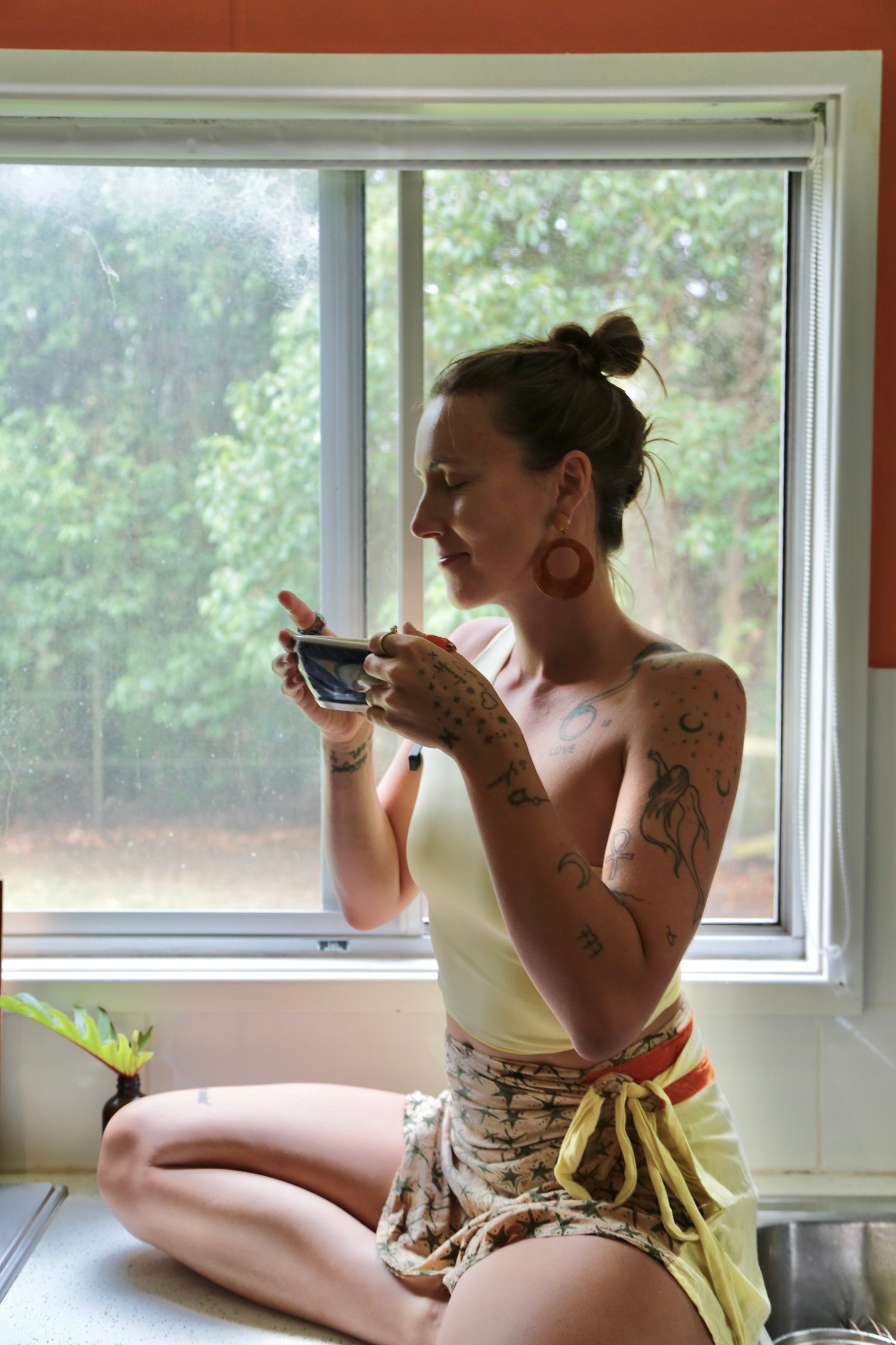 Woman sitting by a window holding a phone, surrounded by greenery outside.