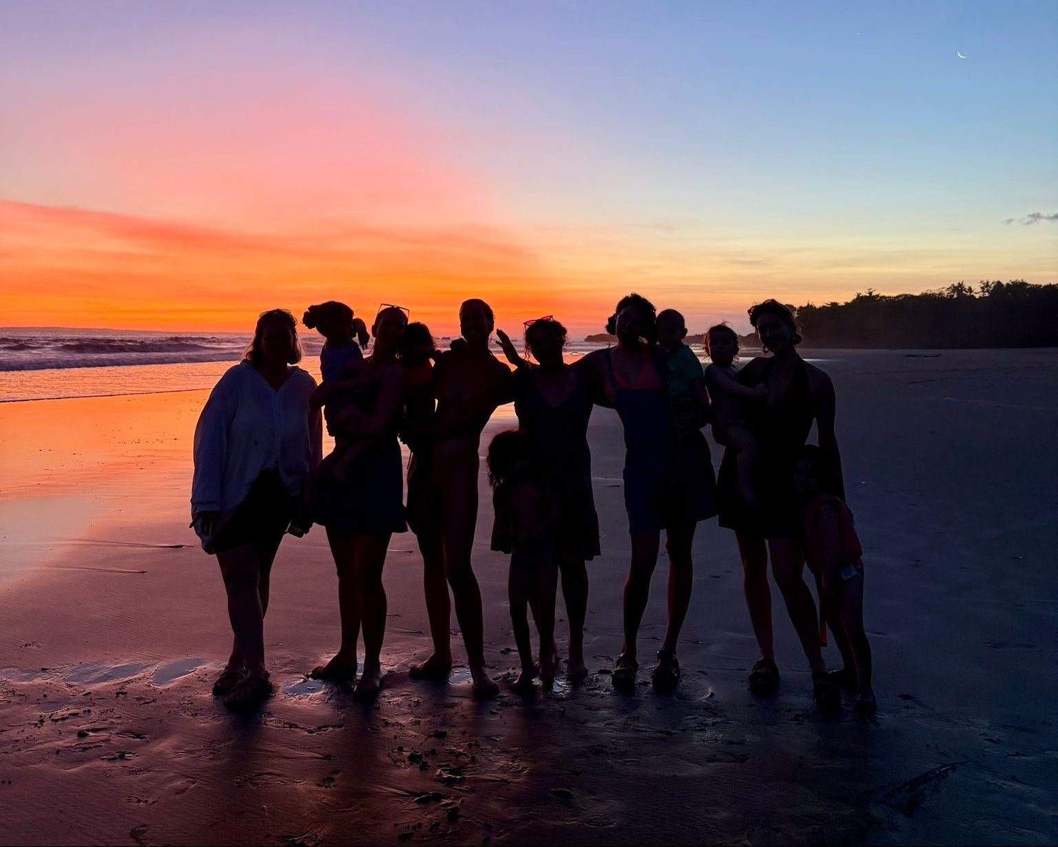 Group of mums standing on a beach at sunset, silhouetted against the colorful sky.